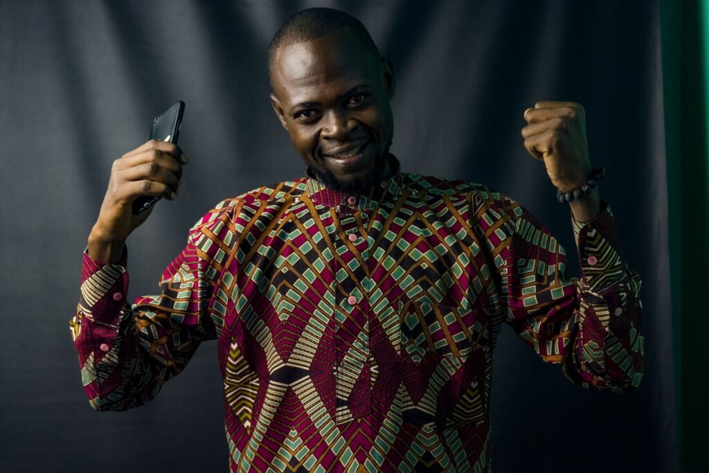 A smiling man in a colourful ankara shirt raises his phone and fist in celebration.