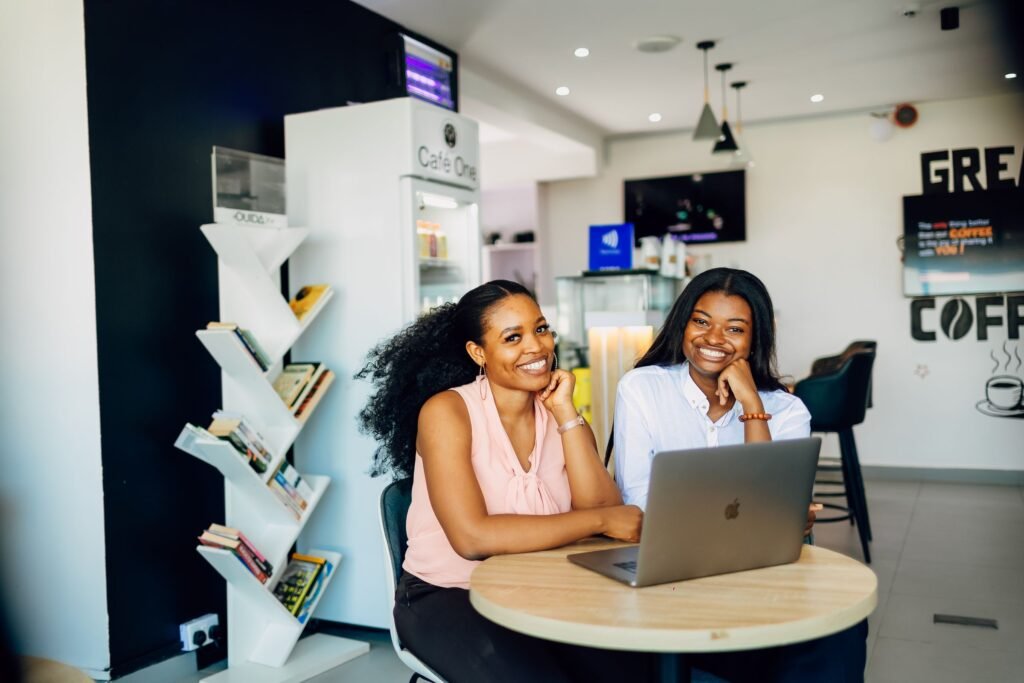 Two smiling women sit together at a laptop in a café in Lagos.