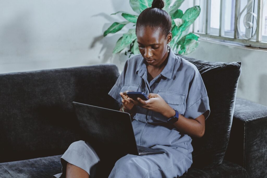 A woman on a sofa in Abuja checks something on her phone with her laptop open.
