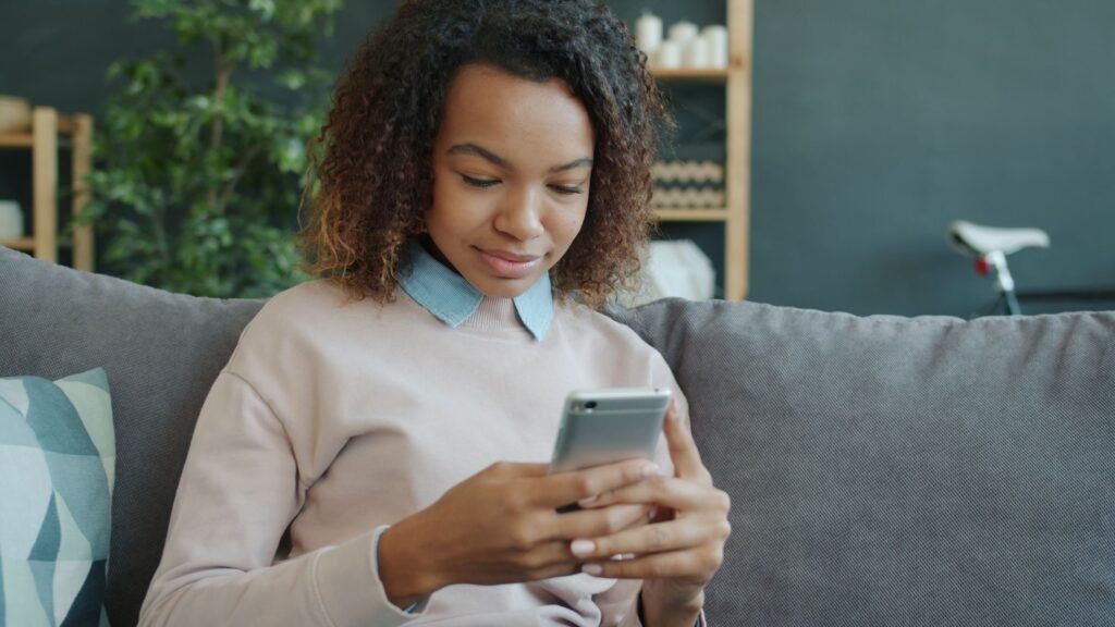 A young woman reads something on her smartphone while sitting on a couch.